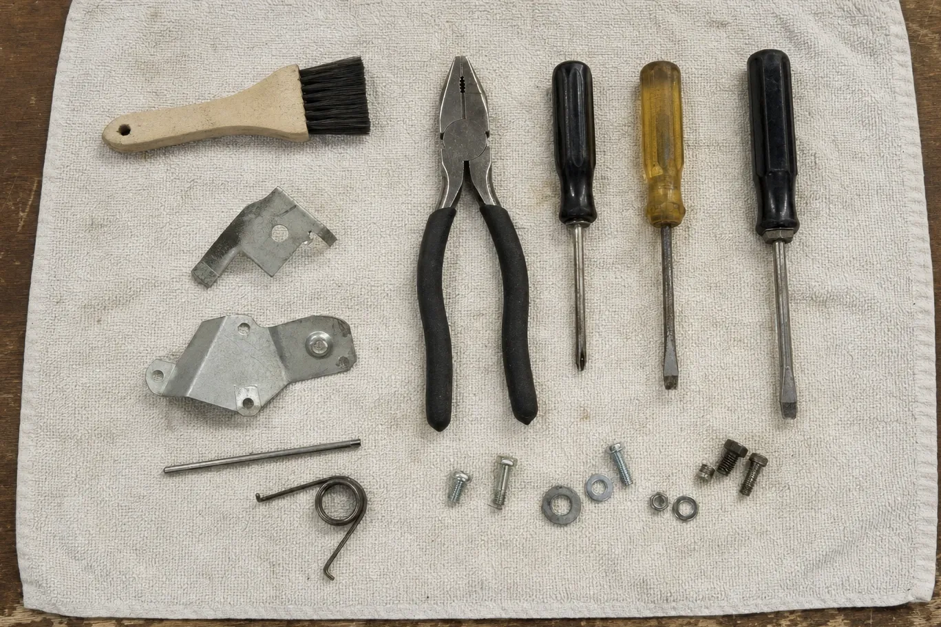 Two galvanised brackets, a torsion spring and a locating pin laid out on a towel beside pliers, three screwdrivers, a bristle brush and rows of screws, washers and nuts